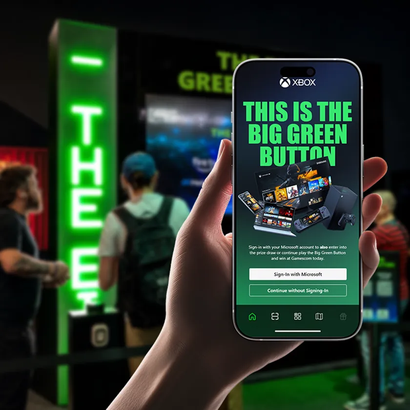 Hand holding a phone showing the Big Green Button web app with sign-in options, in front of an illuminated gaming stand