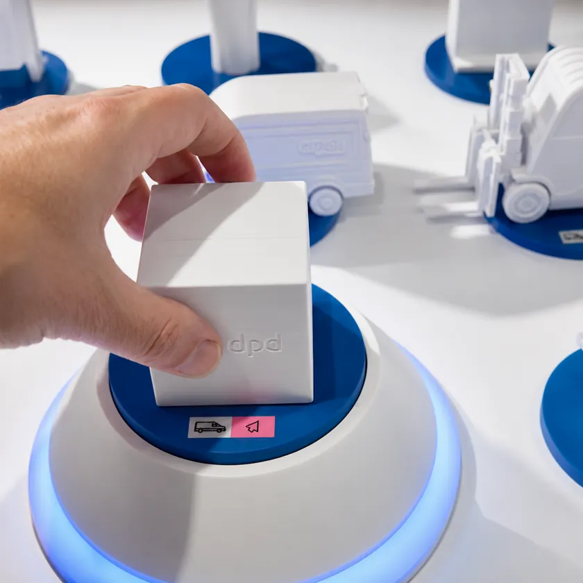 Hand lifting a white 3D-printed parcel model from an illuminated plinth, with model vans and a forklift on blue bases behind