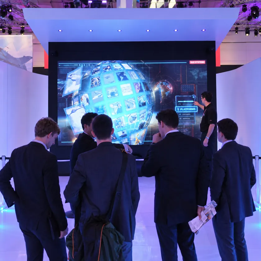 Presenter using a large touchscreen showing a 3D sphere of image tiles and a globe, watched by a group of visitors on an exhibition stand