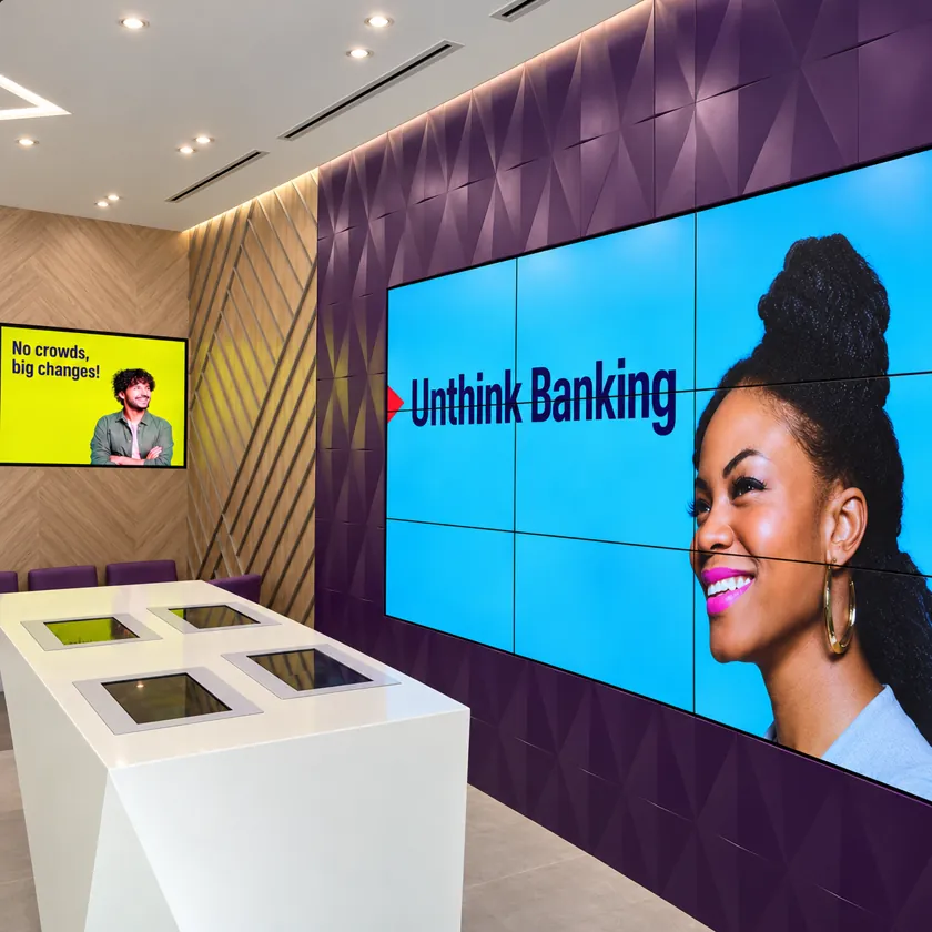 Bank branch interior with a large video wall showing a smiling woman and a campaign line, a digital poster screen on the wood-panelled wall, and a white table with four inset touchscreens